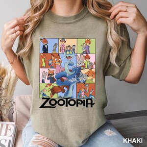 May include: Khaki-colored t-shirt featuring a colorful graphic from the animated film "Zootopia." The design includes various characters in a grid pattern, with the film's title in bold black letters at the bottom. The shirt is a casual wear item.
