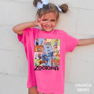 May include: A bright pink t-shirt featuring a colorful graphic from the movie "Zootopia." The design includes various characters from the film, with the word "ZOOTOPIA" in a black font at the bottom. The shirt is a vibrant shade of pink.