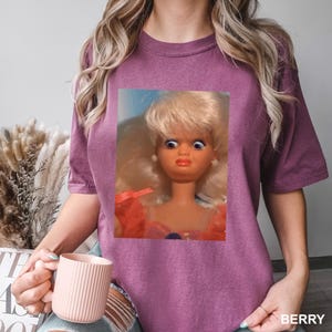 May include: Berry-colored t-shirt featuring a close-up of a vintage doll with a surprised expression. The doll has blonde hair, blue eyes, and is wearing a pink dress. The word "BERRY" is printed in white at the bottom right.