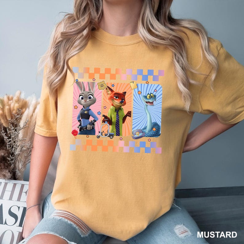 May include: A mustard yellow short-sleeve t-shirt featuring a graphic print of three animated characters: a rabbit in a police uniform, a fox in a green shirt and tie, and a blue snake. The design includes a checkered border and small star icons.