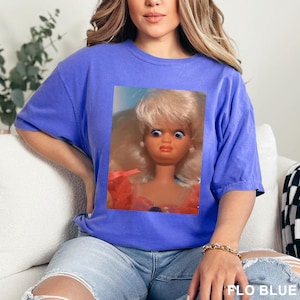 May include: A blue t-shirt featuring a close-up of a vintage doll with blonde hair and wide eyes. The doll is wearing a pink and orange dress. The text "FLO BLUE" is printed at the bottom right of the image.