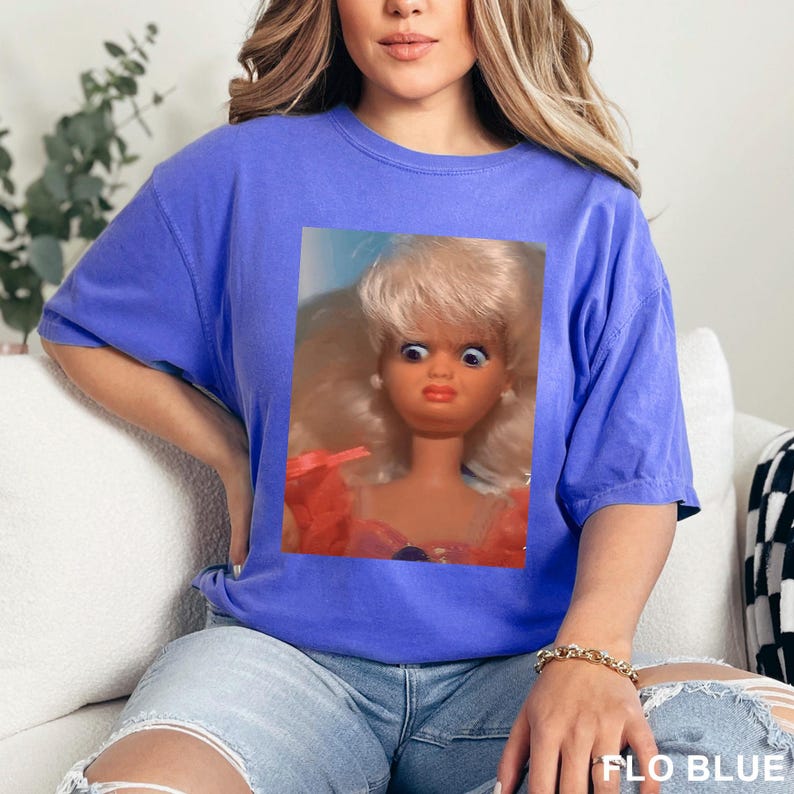 May include: A blue t-shirt featuring a close-up of a vintage doll with blonde hair and wide eyes. The doll is wearing a pink and orange dress. The text "FLO BLUE" is printed at the bottom right of the image.