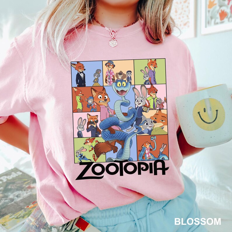 May include: A light pink sweatshirt featuring a colorful graphic of characters from the movie "Zootopia". The graphic is in a grid pattern and includes the word "Zootopia" in black. A white mug with a yellow smiley face is held in the hand.