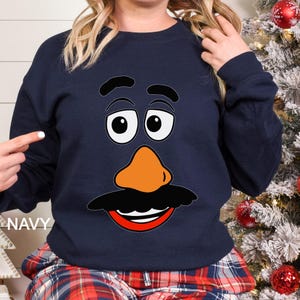 May include: Navy blue sweatshirt featuring a cartoon face with large white-rimmed eyes, an orange nose, a black mustache, and a red smiling mouth. The word "NAVY" is printed on the left side of the sweatshirt.