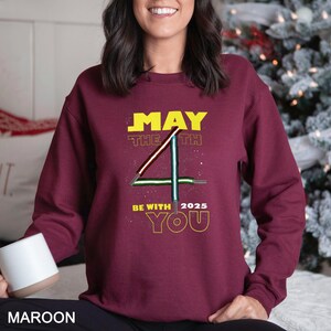 May include: A maroon sweatshirt featuring the text "MAY THE 4TH BE WITH YOU 2025" in yellow. The number 4 is designed with lightsabers. The word "MAROON" is at the bottom of the sweatshirt.