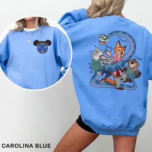 Op de afbeelding: Carolina blauwe sweatshirt met cartoonontwerpen op de voor- en achterkant. De voorkant heeft een karakterontwerp en de achterkant toont een groep geanimeerde personages. De sweatshirt heeft een effen Carolina blauwe kleur.