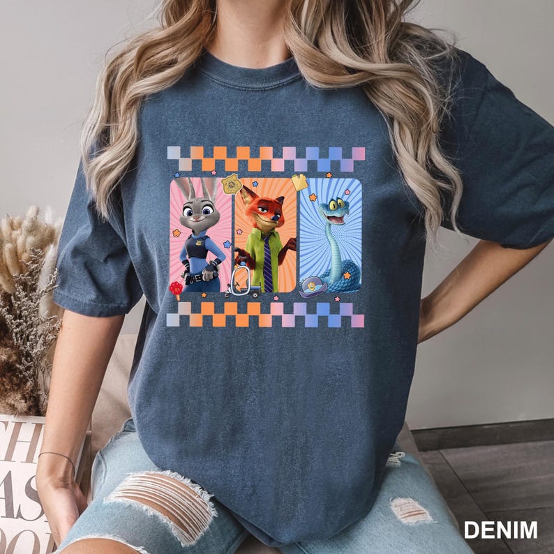 May include: A dark blue, short-sleeved t-shirt featuring a graphic print of three animated characters: a rabbit in a police uniform, a fox in a green shirt and tie, and a blue snake. The design includes a checkered border and small star icons.