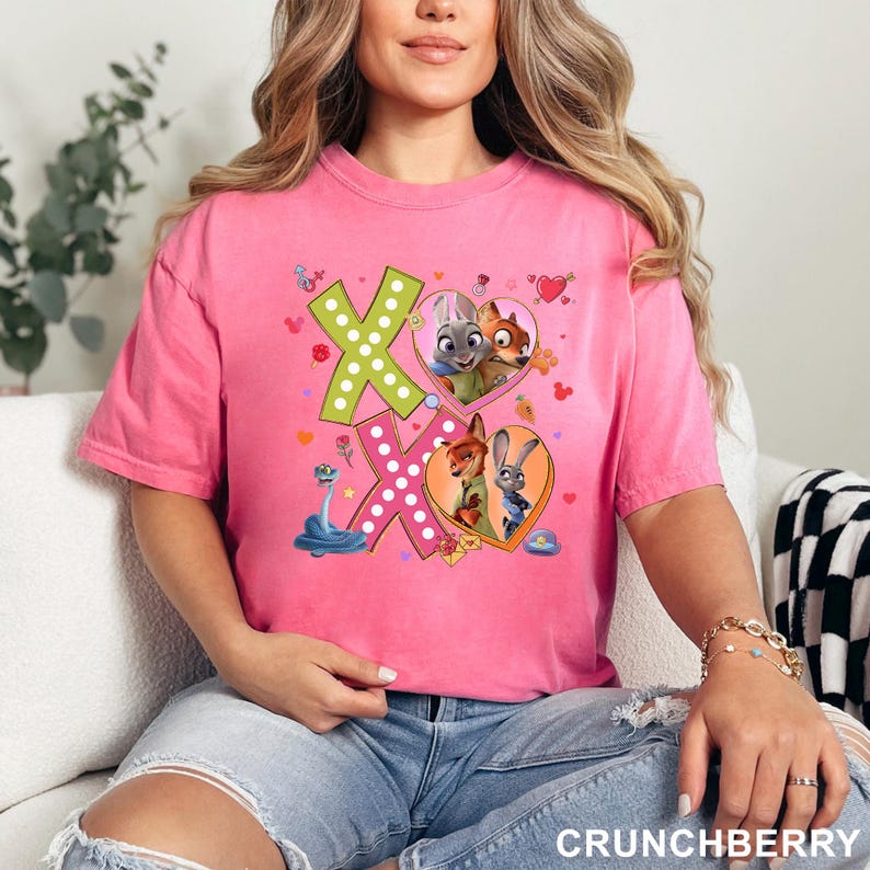 May include: A pink t-shirt featuring a graphic design with the letters "XO" in green and pink, decorated with hearts and cartoon characters. The shirt has short sleeves and a crew neck.