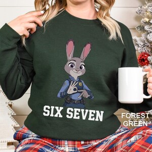 May include: Forest green sweatshirt featuring a cartoon image of Judy Hopps from Zootopia in a police uniform. The text "SIX SEVEN" is printed in white across the front. A white mug is held in the right hand.