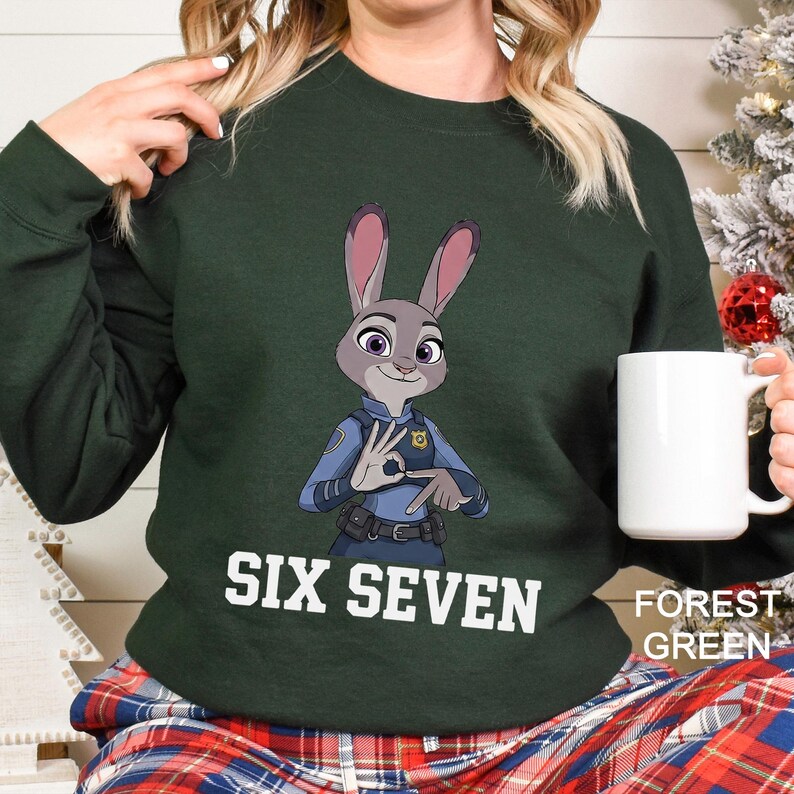 May include: Forest green sweatshirt featuring a cartoon image of Judy Hopps from Zootopia in a police uniform. The text "SIX SEVEN" is printed in white across the front. A white mug is held in the right hand.