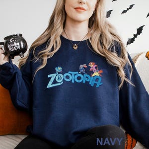 Puede incluir: Sudadera azul marino con la palabra "Zootopia" en color turquesa y personajes de dibujos animados. La sudadera tiene cuello redondo y mangas largas. La palabra "NAVY" est&aacute; impresa en blanco en la parte inferior de la sudadera.