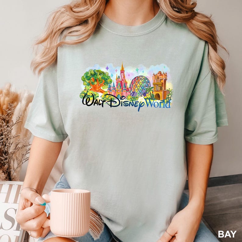 K&ouml;nnte beinhalten: Ein helles salbeigr&uuml;nes Kurzarm-T-Shirt mit einem farbenfrohen Grafikdruck der Walt Disney World-Wahrzeichen, darunter das Schloss, die geod&auml;tische Kuppel und der Baum des Lebens, mit dem Schriftzug Walt Disney World darunter.