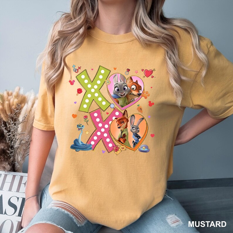 May include: Mustard-colored t-shirt featuring a graphic of the characters from Zootopia. The design includes the letters "XO" with heart-shaped frames containing the characters Judy Hopps and Nick Wilde. The shirt is made of a soft material.