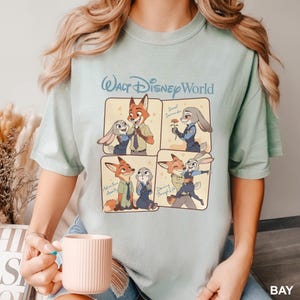 K&ouml;nnte beinhalten: Ein hellgr&uuml;nes T-Shirt mit Rundhalsausschnitt und einem Walt Disney World-Aufdruck, der in vier Feldern die Zeichentrickfiguren Nick Wilde und Judy Hopps in verschiedenen Posen zeigt, darunter mit einer Blume und beim Tanzen, inklusive Text.