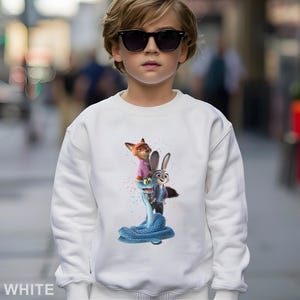 K&ouml;nnte beinhalten: Wei&szlig;es Sweatshirt mit einer farbenfrohen Grafik von Charakteren aus dem Animationsfilm Zootopia. Das Design zeigt einen Fuchs in einem rosa Hemd, ein Kaninchen und eine blaue Schlange. Das Wort "WHITE" ist unten aufgedruckt.