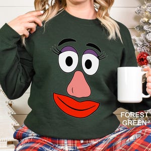 May include: Forest green sweatshirt featuring a cartoon face with large eyes, a pink nose, and red lips. The sweatshirt has a crew neck and long sleeves. The text "FOREST GREEN" is printed on the bottom right corner.