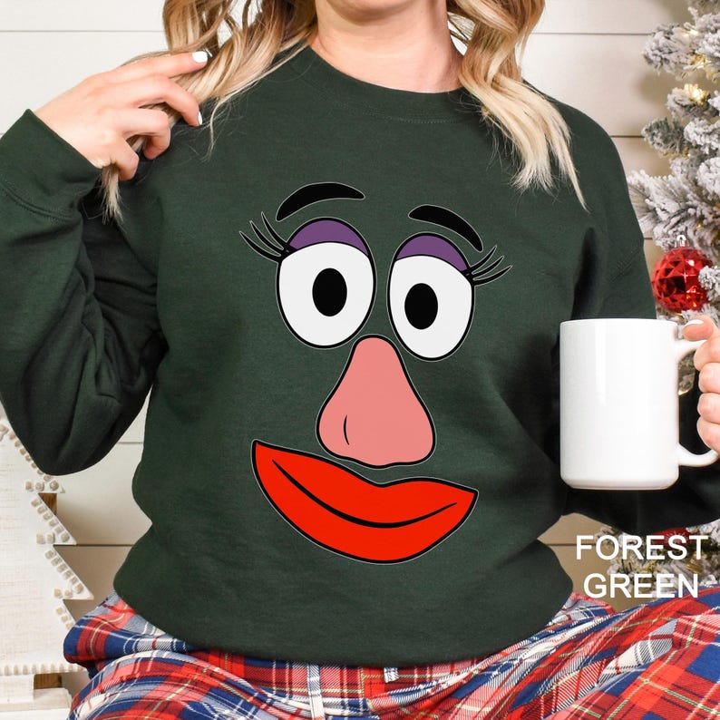 May include: Forest green sweatshirt featuring a cartoon face with large eyes, a pink nose, and red lips. The sweatshirt has a crew neck and long sleeves. The text "FOREST GREEN" is printed on the bottom right corner.