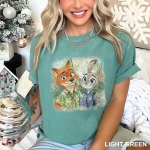 May include: A light green short-sleeve t-shirt featuring a graphic print of a cartoon fox wearing a palm tree patterned shirt and a cartoon rabbit wearing a blue police uniform, both standing together against a textured, speckled background.