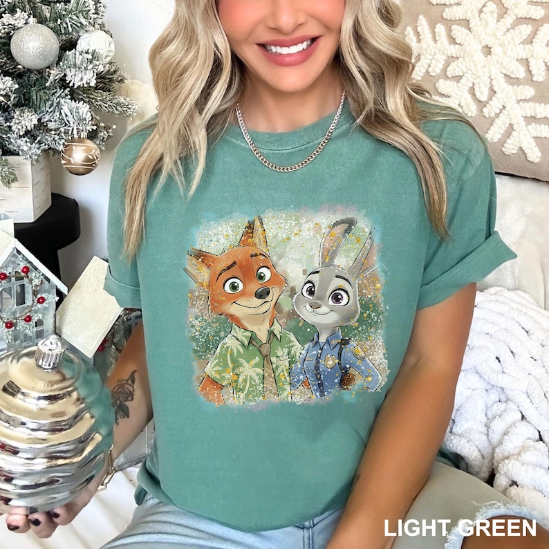 May include: A light green short-sleeve t-shirt featuring a graphic print of a cartoon fox wearing a palm tree patterned shirt and a cartoon rabbit wearing a blue police uniform, both standing together against a textured, speckled background.