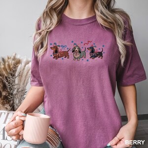 Peut inclure: T-shirt de couleur baie avec trois teckels portant des n&oelig;uds papillon et des chapeaux patriotiques, entour&eacute;s d'&eacute;toiles bleues. Le t-shirt a une coupe d&eacute;contract&eacute;e et des manches courtes. Le mot "BERRY" est imprim&eacute; en bas &agrave; droite.