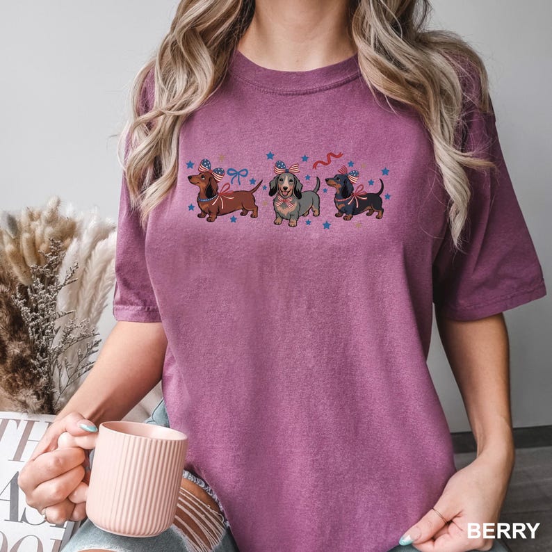 Peut inclure: T-shirt de couleur baie avec trois teckels portant des n&oelig;uds papillon et des chapeaux patriotiques, entour&eacute;s d'&eacute;toiles bleues. Le t-shirt a une coupe d&eacute;contract&eacute;e et des manches courtes. Le mot "BERRY" est imprim&eacute; en bas &agrave; droite.