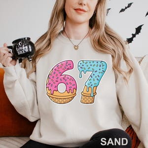 May include: A cream-colored sweatshirt with a colorful graphic of the number 67. The number 6 is pink with sprinkles, and the number 7 is blue with sprinkles. Both numbers are designed to look like ice cream cones.