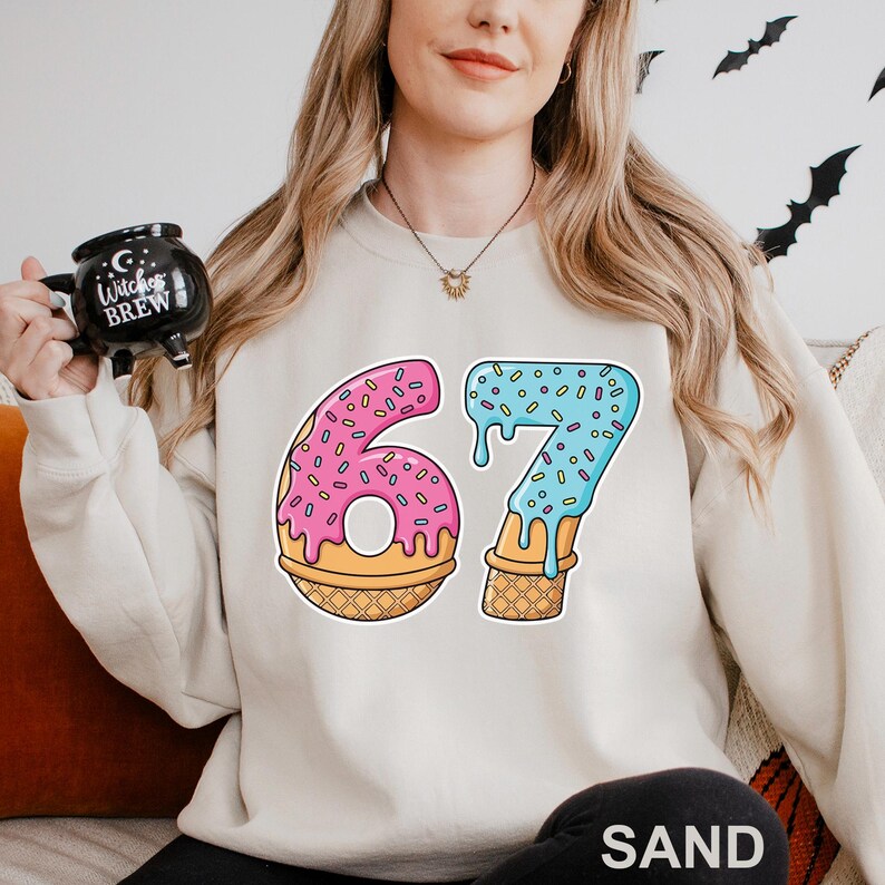 May include: A cream-colored sweatshirt with a colorful graphic of the number 67. The number 6 is pink with sprinkles, and the number 7 is blue with sprinkles. Both numbers are designed to look like ice cream cones.