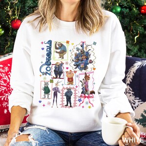 Puede incluir: Sudadera blanca con un gr&aacute;fico colorido de personajes de la pel&iacute;cula animada "Zootr&oacute;polis". El dise&ntilde;o incluye la palabra "Zootr&oacute;polis" en azul, junto con varios personajes de animales en una disposici&oacute;n similar a una cuadr&iacute;cula. La sudadera es de cuello redondo y manga larga.