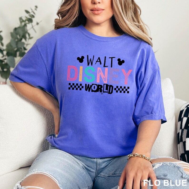 May include: A periwinkle blue short-sleeve t-shirt featuring the text WALT DISNEY WORLD in black and multicolored block letters. The design includes two small black Mickey Mouse head silhouettes and a black and white checkered pattern border.
