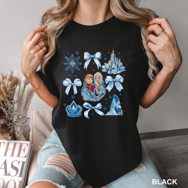 May include: Black t-shirt featuring a colorful graphic of characters, snowflakes, bows, and a castle. The graphic includes the text "BLACK" at the bottom right. The design is in shades of blue, white, and pink.