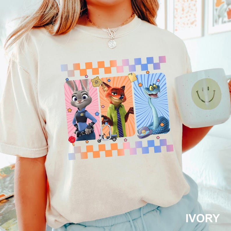 May include: An ivory t-shirt featuring a graphic print of three Zootopia characters: Judy Hopps, Nick Wilde, and a snake. The design includes a checkered border in orange and blue, small star accents, and a police badge icon. The shirt is worn with a light blue bottom.