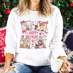 May include: White sweatshirt with a colorful Valentine's Day graphic. The design features cartoon characters in framed portraits, hearts, and the text "Happy Valentine's Day". The sweatshirt is a long-sleeved crew neck.