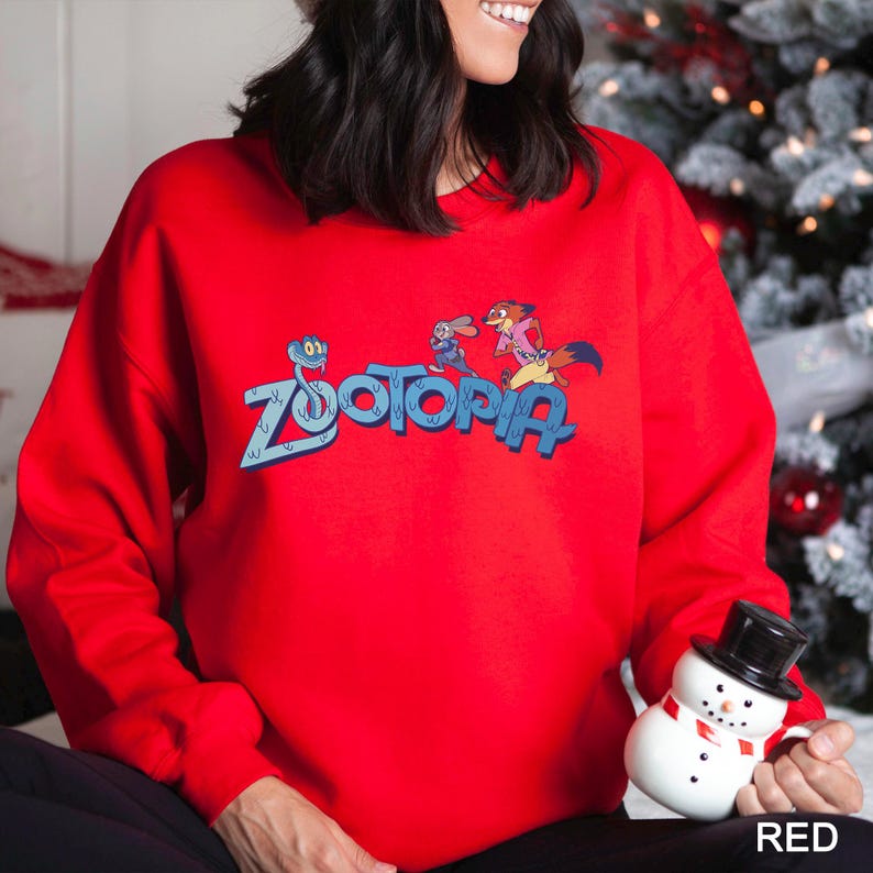 Puede incluir: Una sudadera roja brillante con la palabra "Zootopia" en azul y personajes de dibujos animados. La sudadera es de color rojo liso. La palabra "RED" est&aacute; en la esquina inferior derecha.
