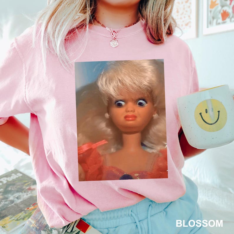 May include: A light pink crew neck t-shirt featuring a doll graphic. The doll has blonde hair and wide eyes. A white mug with a yellow smiley face is held in the right hand. The word "BLOSSOM" is visible in the bottom right corner.
