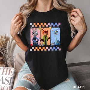 May include: A black t-shirt featuring a graphic print of three animated characters from Zootopia: Judy Hopps, Nick Wilde, and a snake. The design is framed by a checkered border in orange and blue, with small star accents and police-themed icons.