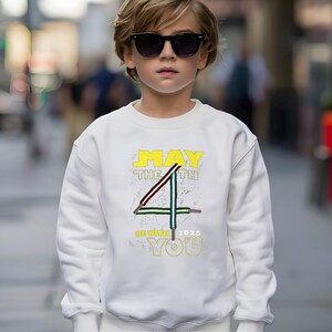 May include: White sweatshirt featuring the text "MAY THE 4TH BE WITH YOU 2025" in yellow and green. The design includes a stylized number 4 with red and green accents. The sweatshirt is worn by a person wearing sunglasses.