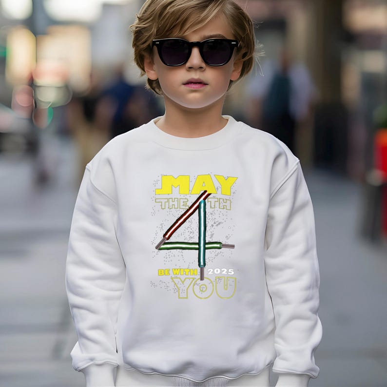 May include: White sweatshirt featuring the text "MAY THE 4TH BE WITH YOU 2025" in yellow and green. The design includes a stylized number 4 with red and green accents. The sweatshirt is worn by a person wearing sunglasses.