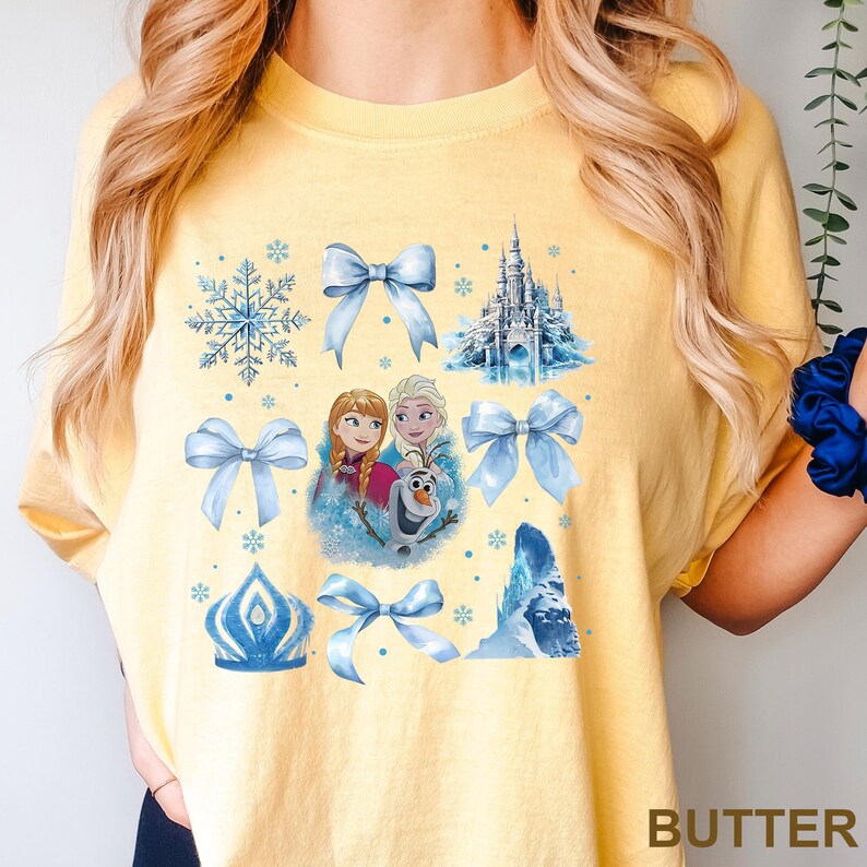 May include: Butter yellow t-shirt featuring a collage of light blue Frozen-themed graphics. The design includes snowflakes, bows, a castle, and images of Anna, Elsa, and Olaf. The word "BUTTER" is printed at the bottom right.