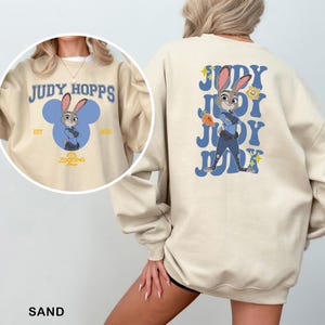 K&ouml;nnte beinhalten: Sandfarbener Sweatshirt mit einem Cartoon-Bild von Judy Hopps aus Zootopia. Die Vorderseite hat ein blaues Mickey-Mouse-f&ouml;rmiges Design mit Judy Hopps und dem Text "Judy Hopps". Die R&uuml;ckseite hat ein gr&ouml;&szlig;eres Bild von Judy Hopps mit dem Text "Judy".