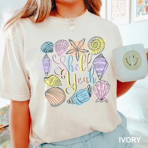 May include: Ivory-colored t-shirt with a colorful seashell graphic and the text "Shell Yeah." The design includes various seashells and a starfish in pastel colors. A white mug with a smiley face is also visible.