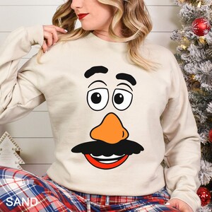 May include: Sand-colored sweatshirt featuring a cartoon face with black eyebrows, large eyes, an orange nose, and a black mustache with a red mouth. The word "SAND" is printed in white at the bottom. The sweatshirt is part of a holiday-themed collection.