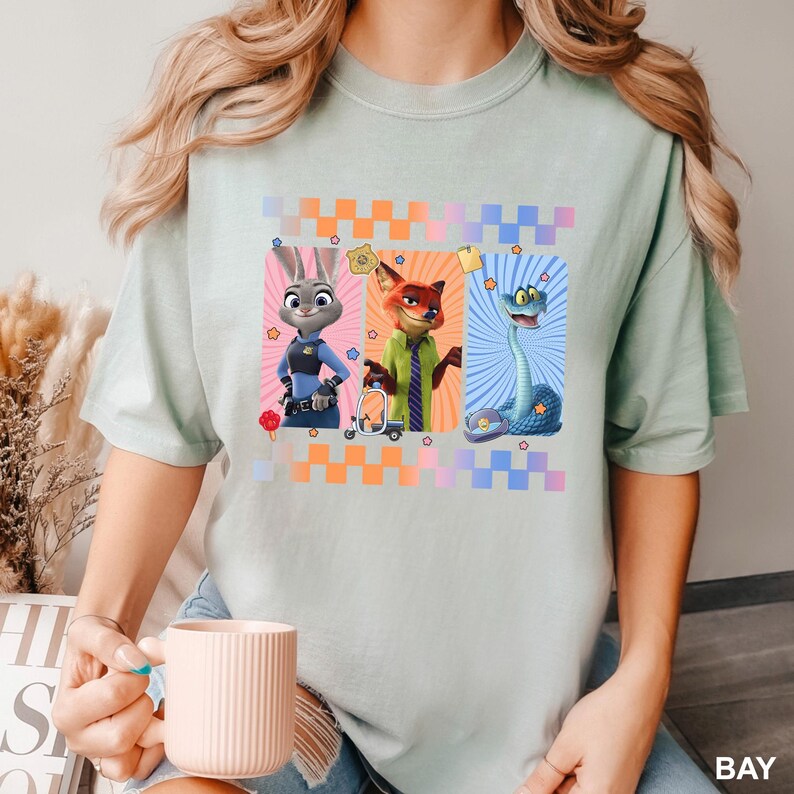 May include: A light green t-shirt featuring a graphic print of three animated characters: a rabbit in a police uniform, a fox in a green shirt and tie, and a blue snake. The design includes a checkered border, small stars, and a badge icon.
