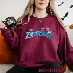 Puede incluir: Sudadera granate con la palabra "Zootopia" en azul y personajes de dibujos animados. La sudadera es de color granate liso. La palabra "MAROON" est&aacute; impresa en la parte inferior de la imagen.