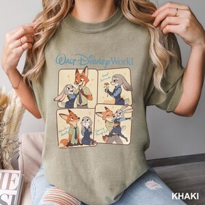 K&ouml;nnte beinhalten: Ein khakifarbenes T-Shirt mit Rundhalsausschnitt und einem Walt Disney World-Grafikdruck, der in vier Feldern die Zeichentrickfiguren Nick Wilde und Judy Hopps in verschiedenen Szenen zeigt, inklusive der Texte Sweet Surrender, Adventure Awaits und Dancing Through Life.