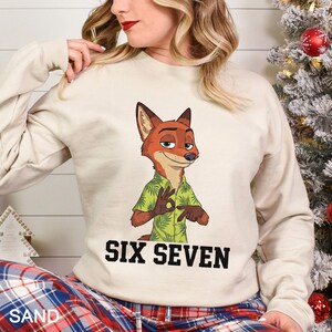 May include: A sand-colored sweatshirt featuring a cartoon fox character in a green Hawaiian shirt, making an 'okay' hand gesture. The text "SIX SEVEN" is printed in bold black letters below the character. The bottom of the sweatshirt is partially visible.