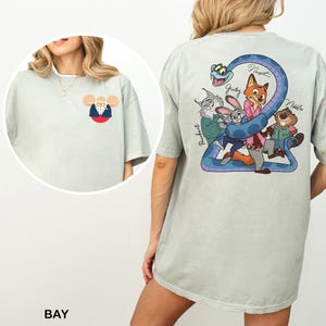 May include: Pale green t-shirt with a cartoon illustration on the back, including a blue snake and various animal characters. The front displays a small cartoon mouse in glasses and a suit. The word "BAY" is printed at the bottom of the image.