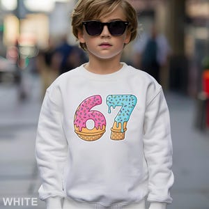 May include: White sweatshirt featuring a colorful graphic of the number 67 designed as ice cream cones. The number 6 is pink with sprinkles, and the number 7 is blue with sprinkles. The sweatshirt is worn by a person wearing sunglasses.