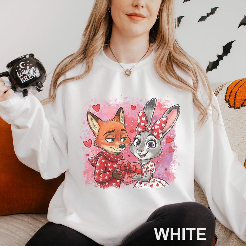 K&ouml;nnte beinhalten: Wei&szlig;es Sweatshirt mit einem Cartoon-Fuchs und -Hasen, die ein herzf&ouml;rmiges Geschenk halten. Der Fuchs tr&auml;gt einen roten Schal, und der Hase hat eine rote Schleife. Das Design umfasst rosa Herzen und das Wort "WHITE" unten.