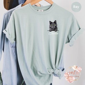 May include: A light blue t-shirt with a gray cat peeking out of a pocket. The cat has green eyes and the text "Charlie" is printed below the cat.