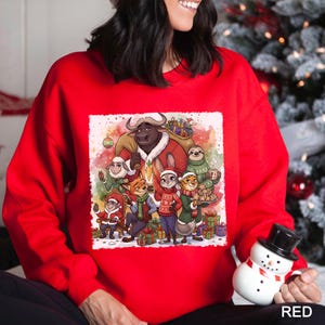 Op de afbeelding: Een rode sweatshirt met een feestelijke illustratie van verschillende dierenfiguren in een winterse sc&egrave;ne. Het ontwerp bevat een stier, een konijn, een vos en andere dieren, allemaal gekleed in feestelijke kleding. Het woord "RED" staat onderaan.